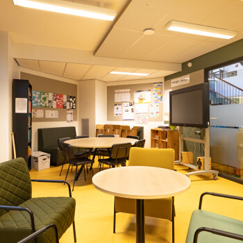 ruimte met een zithoek met 4 fautuils en een ronde tafel met stoelen. een kast en een zitbankje. De vloer is geel van kleur.