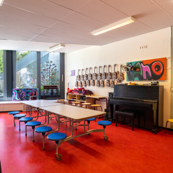 lokaal met veel daglicht, opklaptafels met vaste krukken. Diverse muziekinstrumenten zoals zwarte piano, aan de wand kindergitaartjes, zilverkleurige trommel. Diverse krukken. Rode vloer.