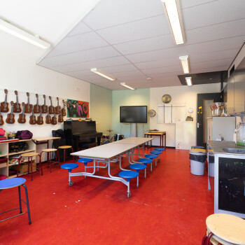 opklaptafels met vaste krukken. Gitaartjes aan de wand links, aan de rechterwand een spoelbak en kast. Aan het einde van het lokaal een digibord. Rode vloer.