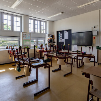 binnenzijde van een klaslokaal met schoolmeubilair, op de schooltafels staan alle stoelen op de kop. 