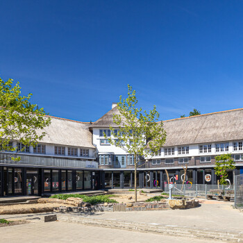 Buitenzijde van de witte school, achterzijde van het pand en is de hoofdingang van de school. Entree in midden van het pand met links en recht een vleugel. Rietgedekte kap. Aan de hemel een stralend blauwe lucht.