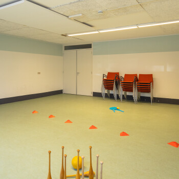 groene sportvloer, witte wanden waartegen enkele oranje stoelen,  kegels en pionnen op de vloer.